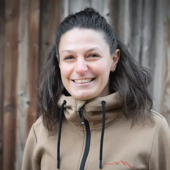 Eine Frau mit gewelltem braunem Haar lächelt, sie steht vor einer Holzwand und trägt eine hellbraune Jacke mit Reißverschluss.