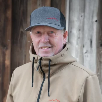 Ein lächelnder Mann in hellbrauner Jacke und grauer Mütze steht vor einer Holzwand.