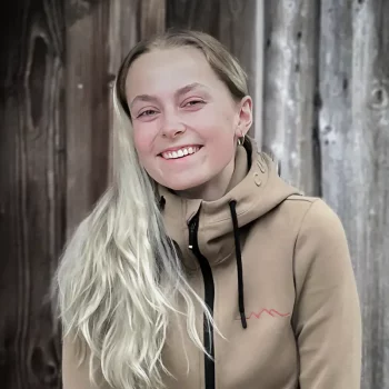 Junge Frau mit langen blonden Haaren, lächelnd, in einem hellbraunen Kapuzenpulli, steht vor einer Holzwand.