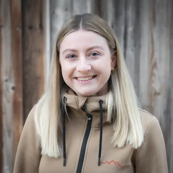 Junge Frau mit glattem blondem Haar, lächelnd, in einer hellbraunen Jacke, steht vor einer Holzwand.