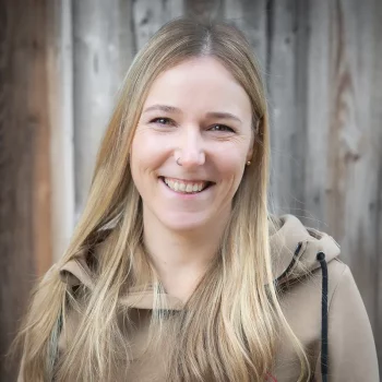 Lächelnde Frau mit langen blonden Haaren und hellbraunem Kapuzenpulli, die vor einem hölzernen Hintergrund steht.