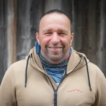 Ein Mann in einem hellbraunen Kapuzenpulli steht vor einer Holzwand und lächelt in die Kamera.